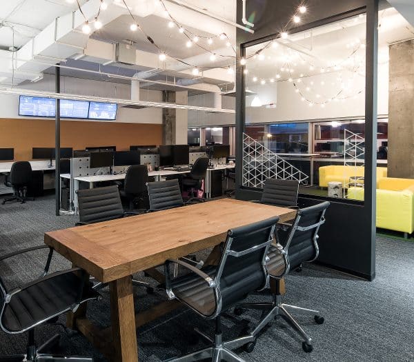 Un bureau moderne avec une longue table en bois entourée de chaises noires, des guirlandes lumineuses et une cloison en verre.
