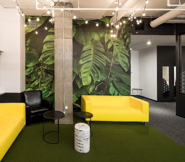 Un canapé jaune et un fauteuil noir dans une pièce avec un mur végétal tropical et un tapis vert.