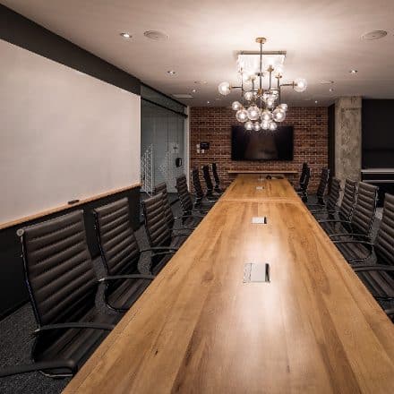 Une grande table de conférence en bois est entourée de chaises noires dans une salle de réunion moderne.