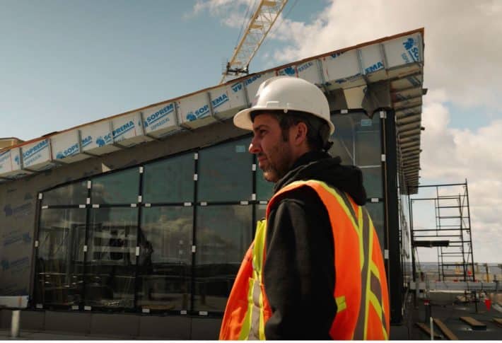 A construction worker in a safety vest and hard hat stands in front of a partially built modern building.