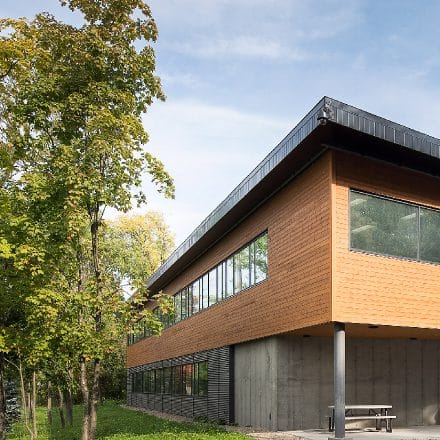 Un bâtiment moderne en bois, avec de grandes fenêtres, se trouve à côté d'arbres feuillus.