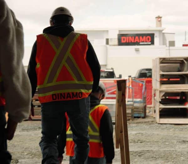 Construction workers wearing high-visibility vests walk past a Dinamo Construction sign.